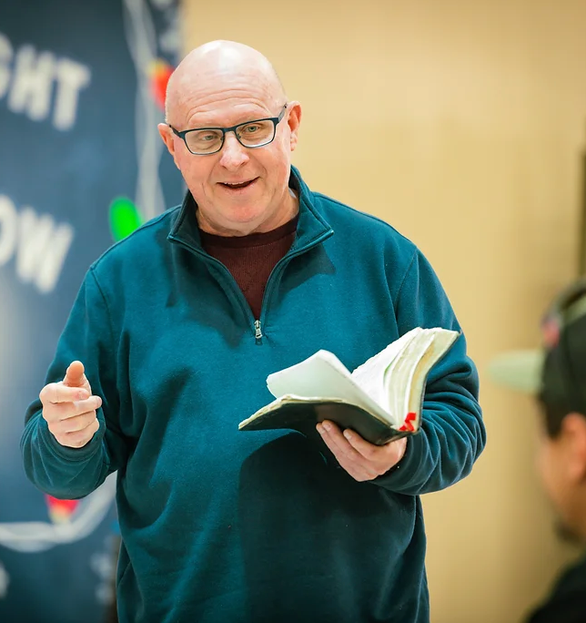 A bald man wearing glasses is holding a book and giving a thumbs up