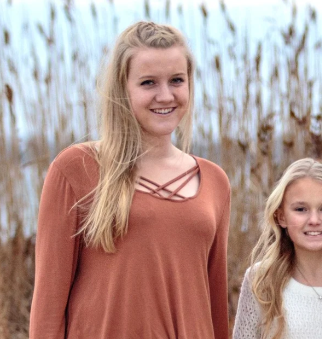Two young women are standing next to each other in front of a field of tall grass.