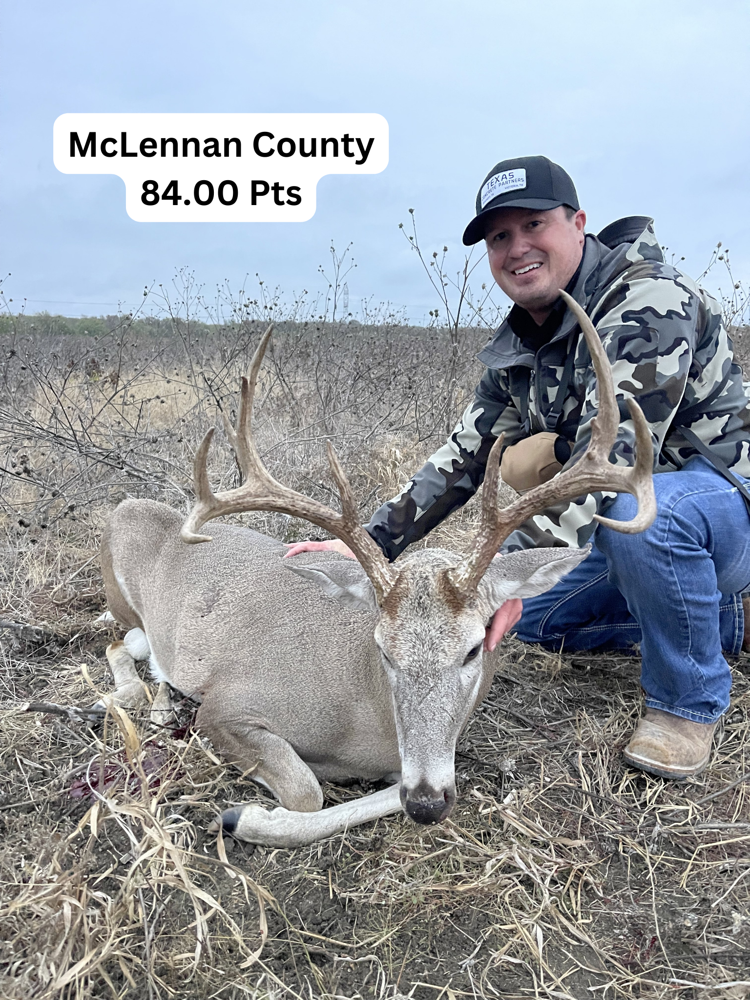 A man kneels beside a harvested deer in a field, McLennan County. The deer has an 84-point rack.