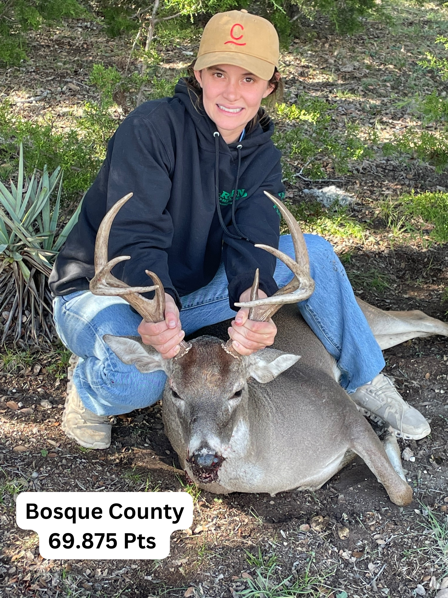 Woman in cap and hoodie kneels beside a harvested deer in Bosque County, Texas.