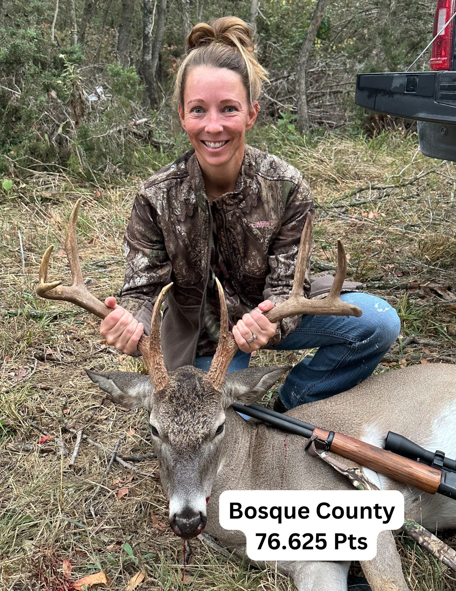 Woman kneels with a deer she shot in Bosque County, Texas, holding its antlers. Smiling in camo.