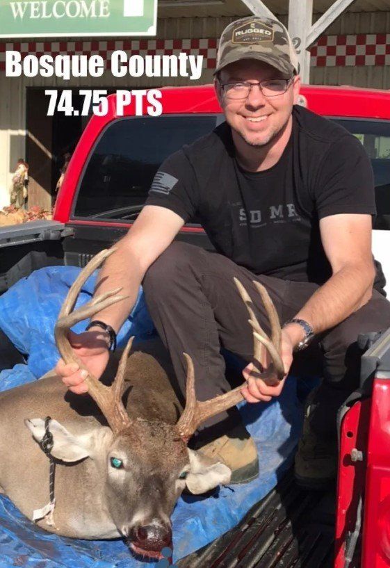 Man in a camouflage hat poses with a deer in Bosque County, Texas. He is smiling and holds the antlers.