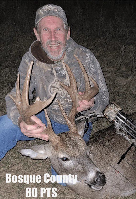 Man in camo holds a large buck's antlers, smiling in Bosque County, 80 points, near a bow.