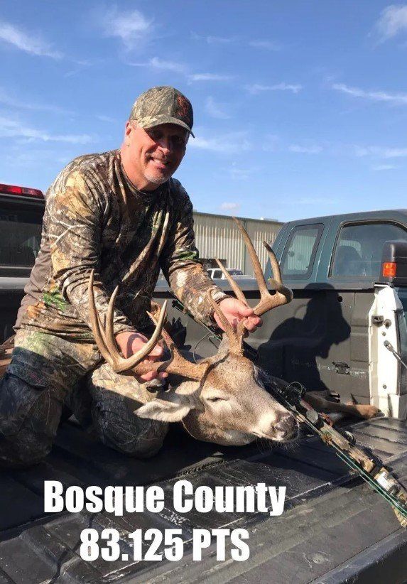 Man kneels with a deer in a truck bed, Bosque County, 83.125 PTS. He smiles, wearing camo, sunny day.