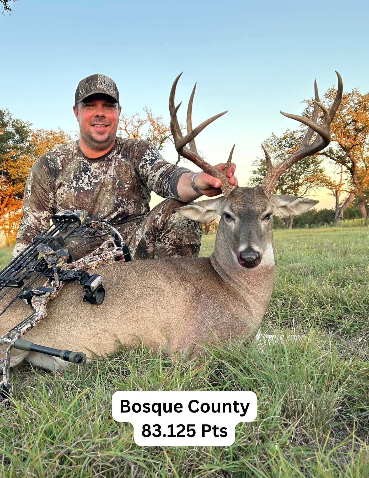 Man in camo with a large buck in Bosque County, Texas. He is smiling and touching the antlers. Grass and sunset in the background.