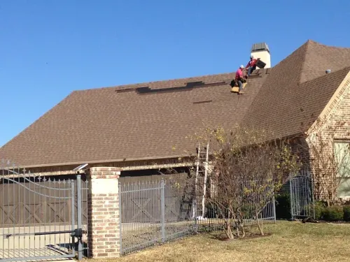Two people on a brown shingle roof, repairing shingles. A ladder is propped against the roof. Sunny day.