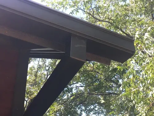 Dark brown rain gutter with a metal support bracket, attached to a brown wooden beam. Trees in background.