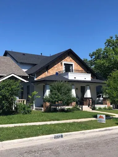 A white house with a black roof, porch, and cedar shake siding. Green lawn and blue sky.