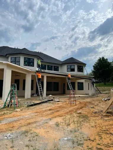 Construction workers on ladders working on a house roof. Cloudy sky, dirt ground.