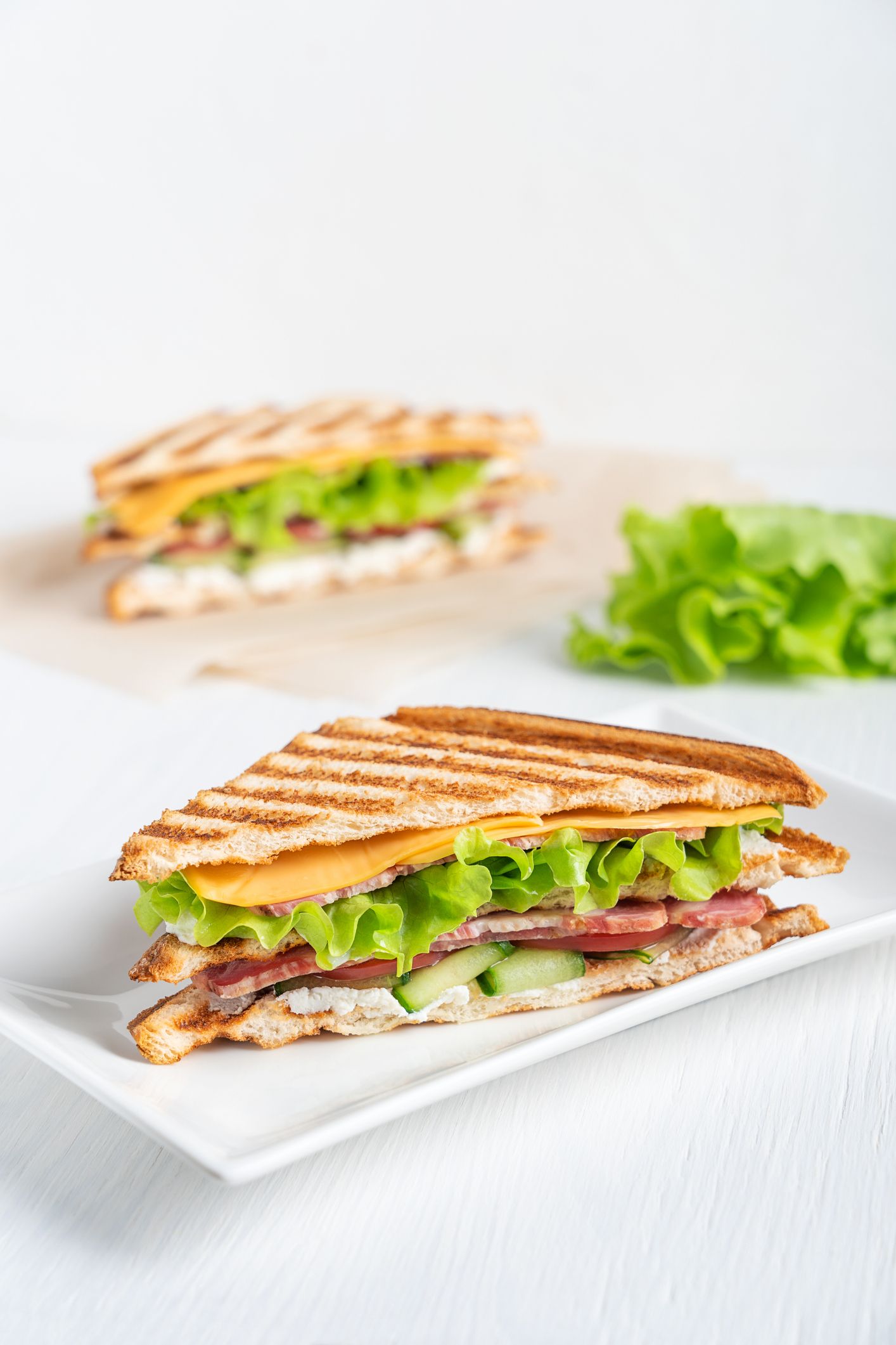 A toasted sandwich with cheese, meat, lettuce, and cucumber on a white plate, with another sandwich in the background.