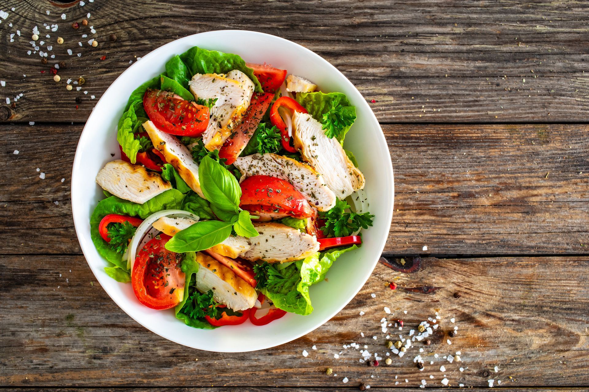 A salad with chicken , lettuce , tomatoes and peppers in a white bowl on a wooden table.