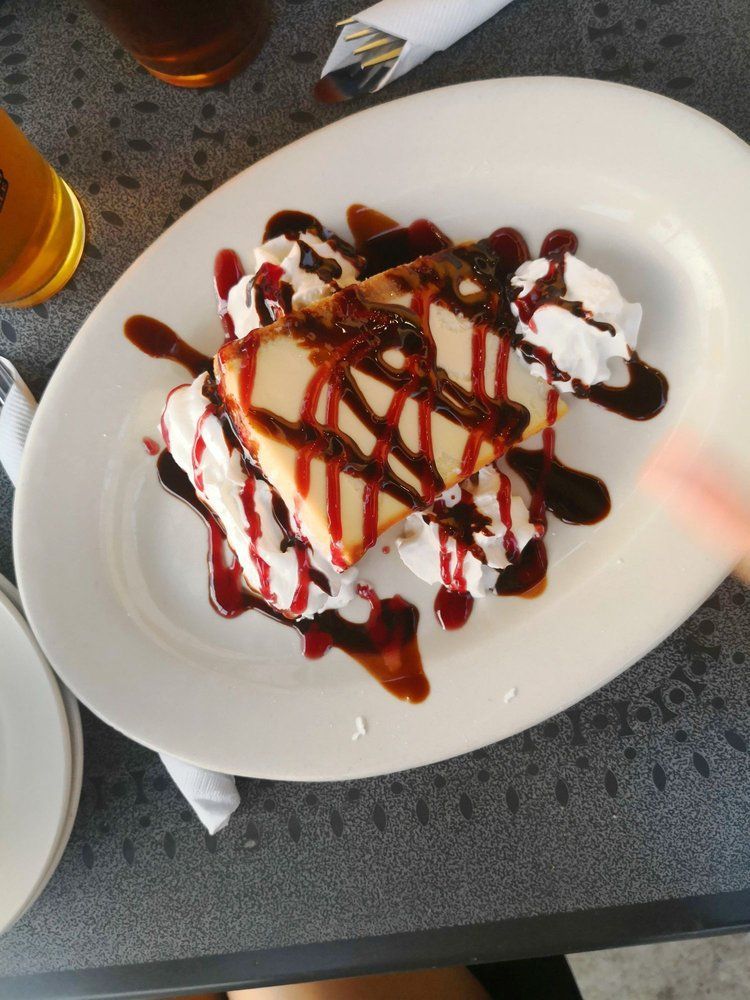 A slice of cheesecake on a white plate, topped with chocolate and raspberry drizzle and swirls of whipped cream.