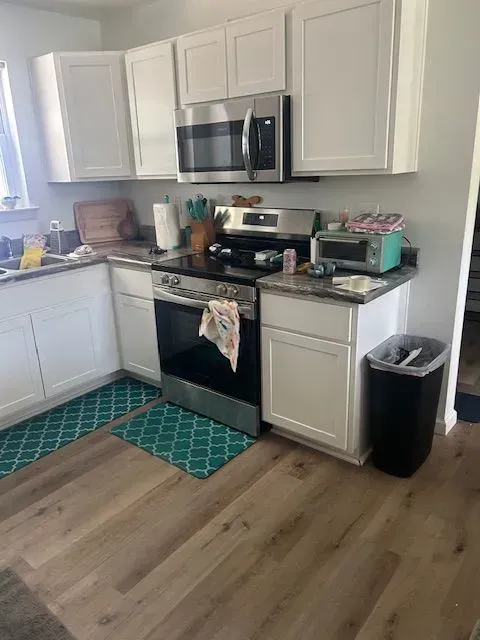 A kitchen with white cabinets , a stove , a microwave , and a trash can.