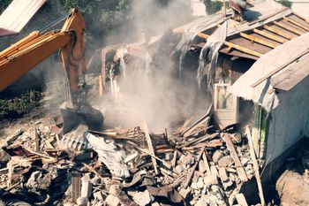 A yellow excavator is demolishing a house with smoke coming out of it.