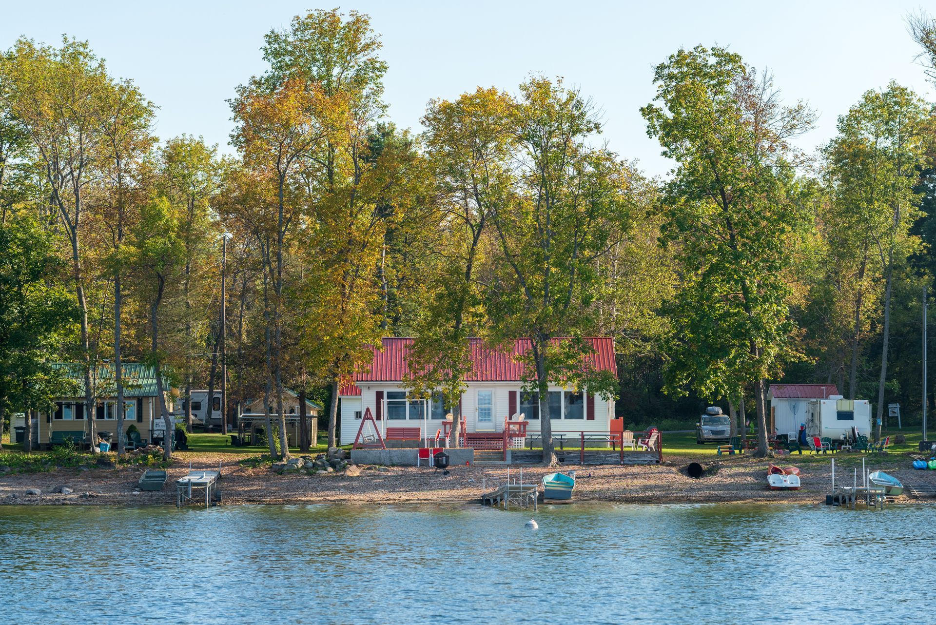 Lakefront cottages with red roofs beneath tall trees, seen across calm water.