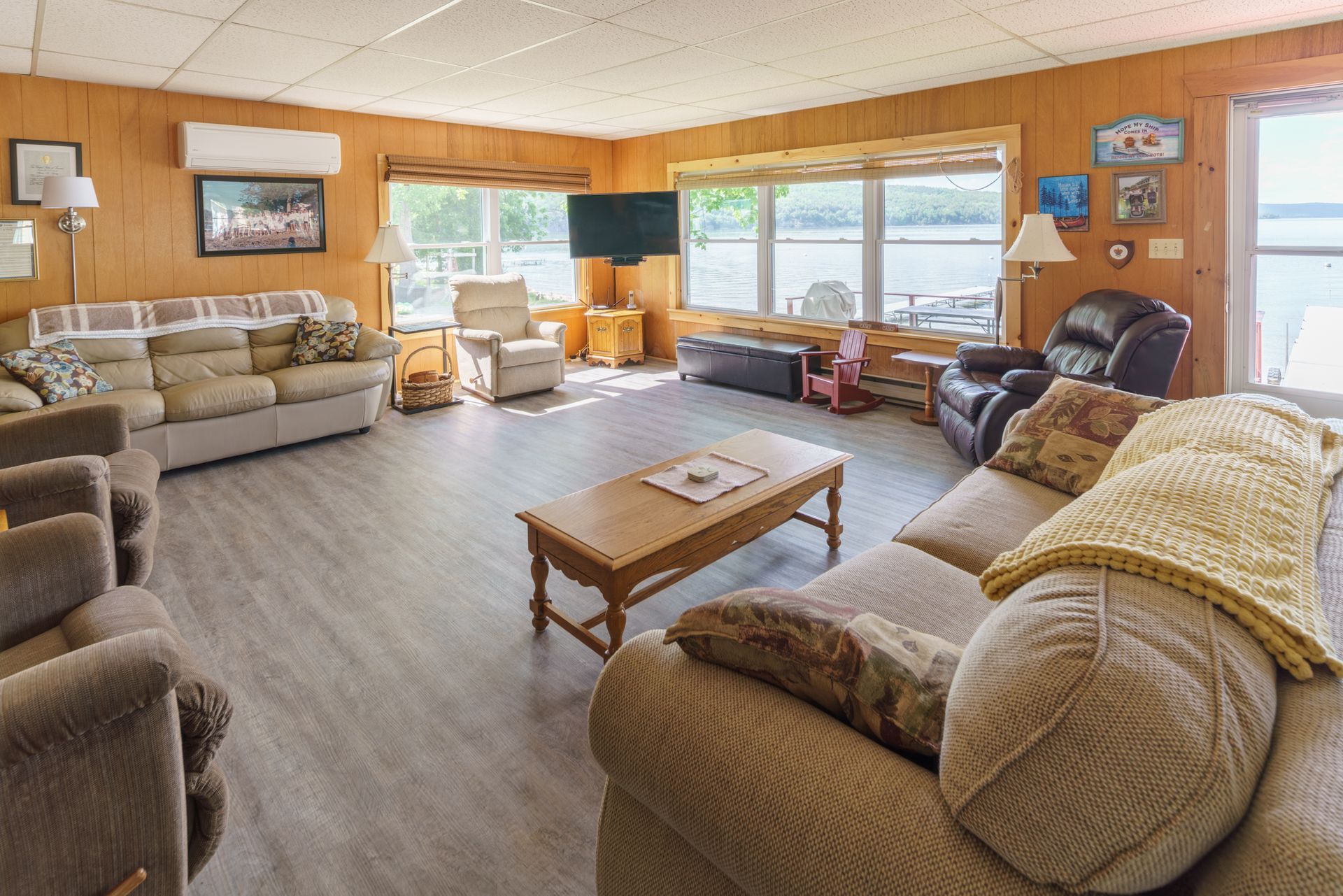 Spacious living room with beige sofas, wood-paneled walls, and large windows overlooking a lake.