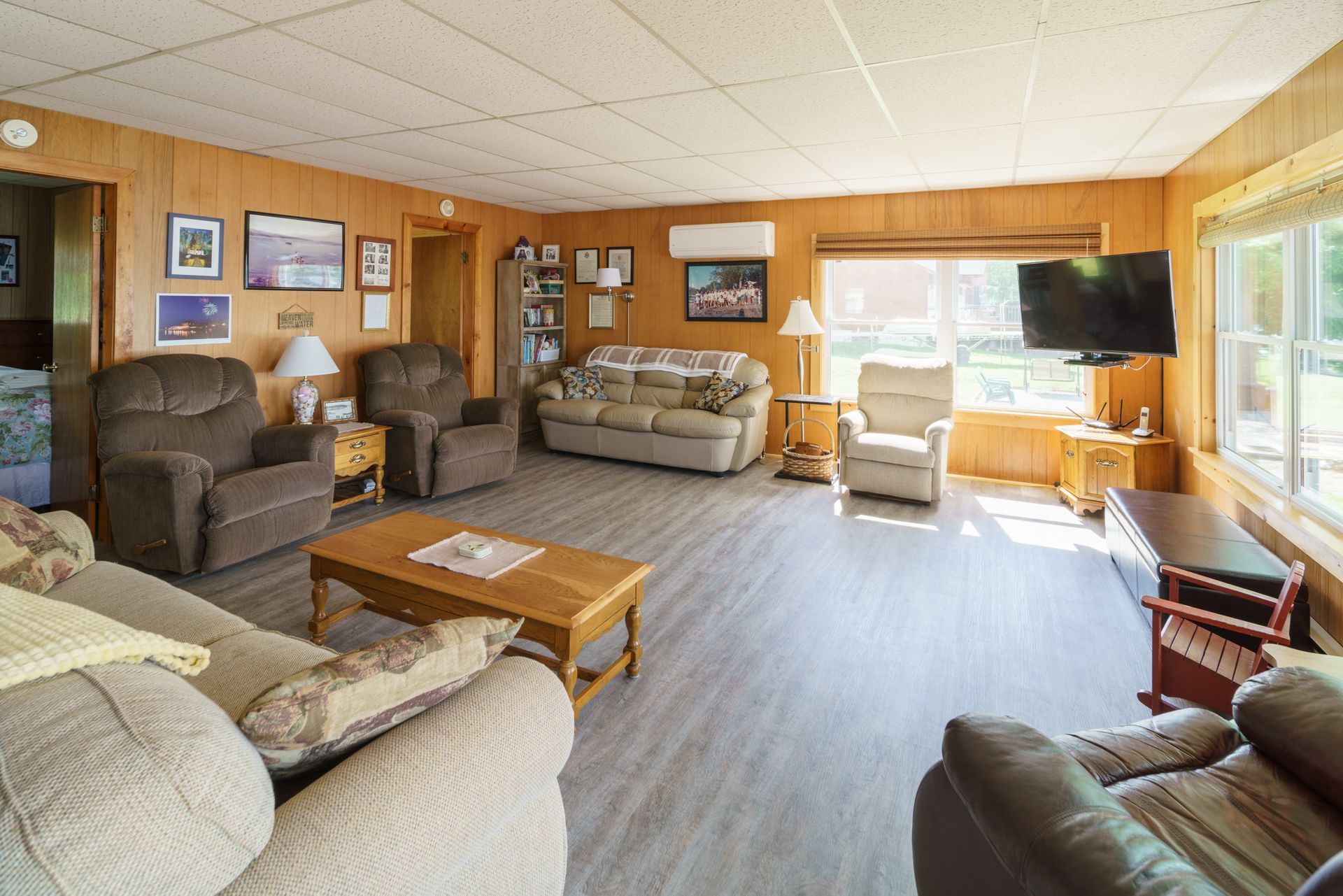 Cozy living room with tan recliners, sofas, a coffee table, wood-paneled walls, and bright windows