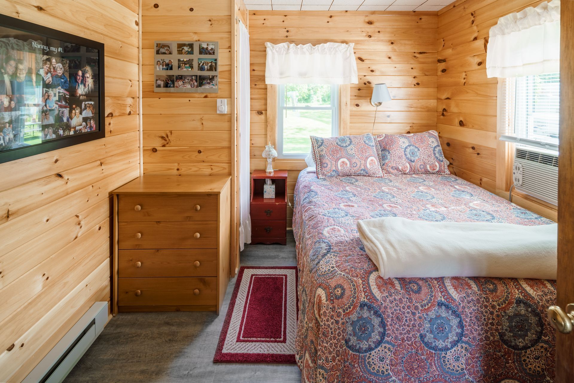 Small cozy wood-paneled bedroom with a patterned bedspread, dresser, windows, and wall-mounted TV.