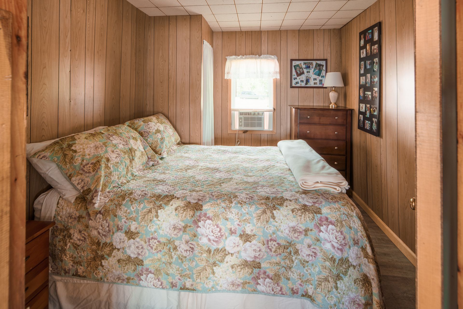 Wood-paneled bedroom with a floral bedspread, dresser, and small window at the back.