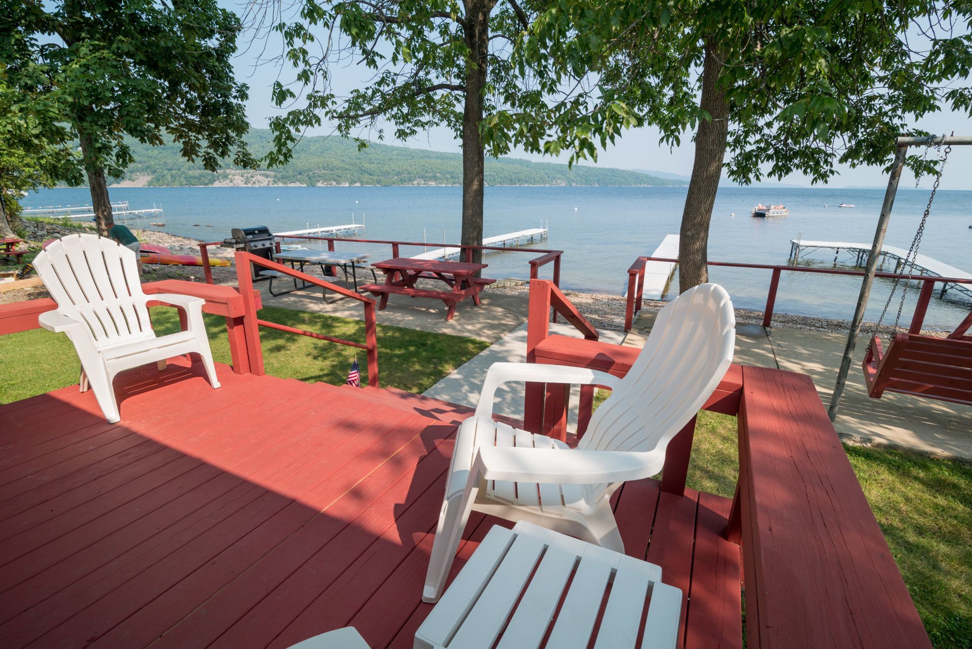 Red deck with white chairs overlooking a lake and dock under leafy trees