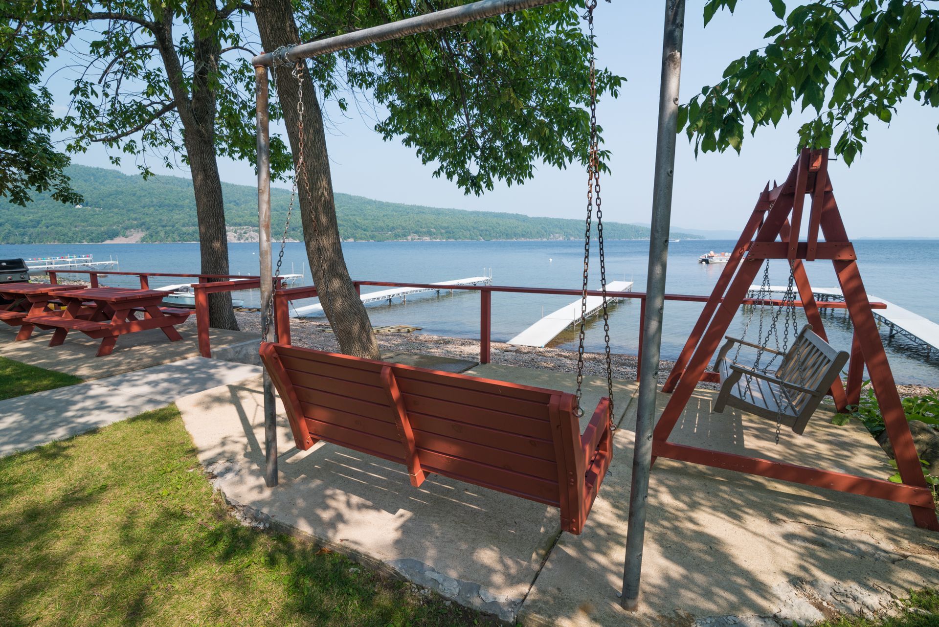 Red wooden swings and benches under trees by a lakeside with picnic tables and a distant shoreline.