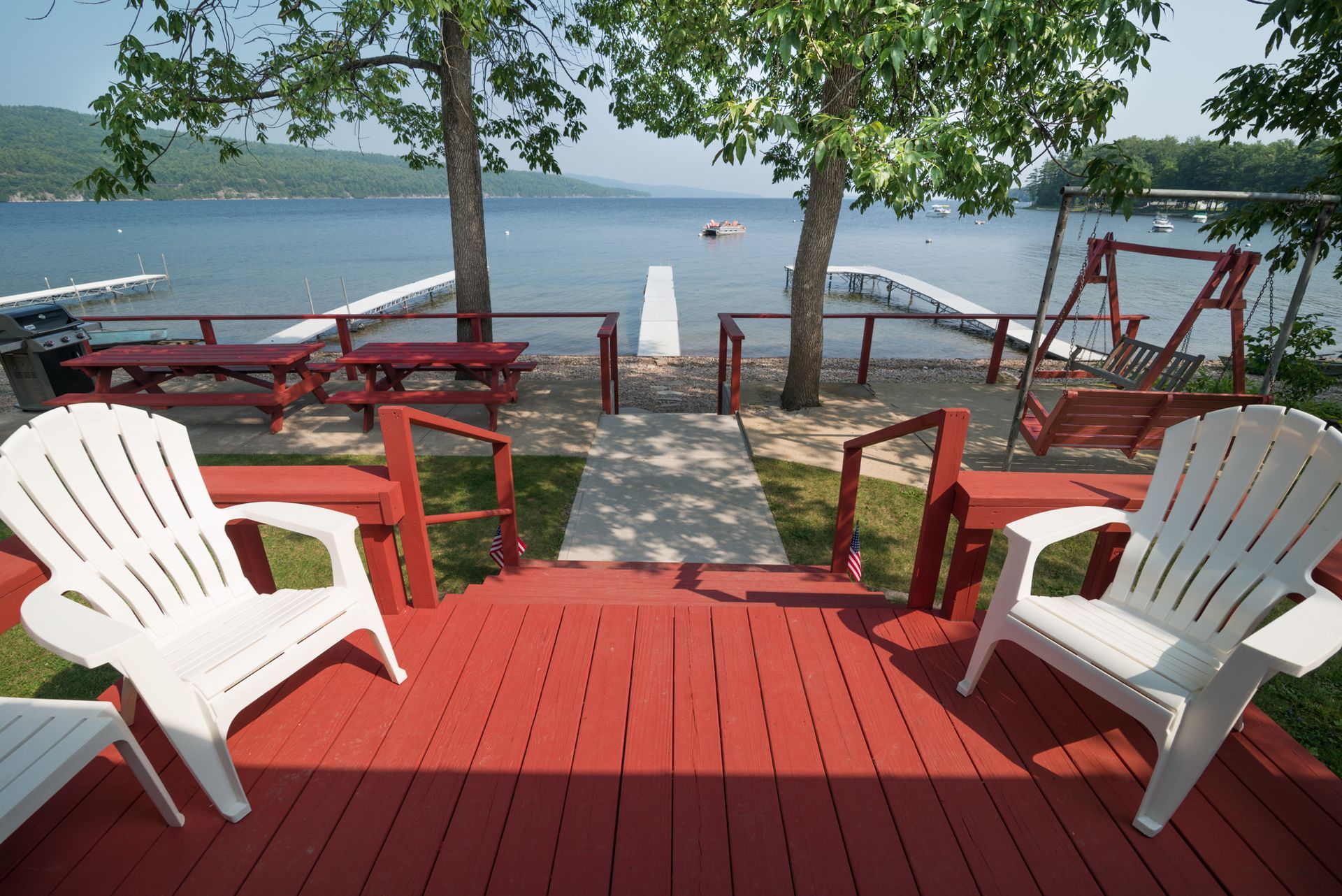 Red deck with white chairs overlooking a lake and dock under leafy trees