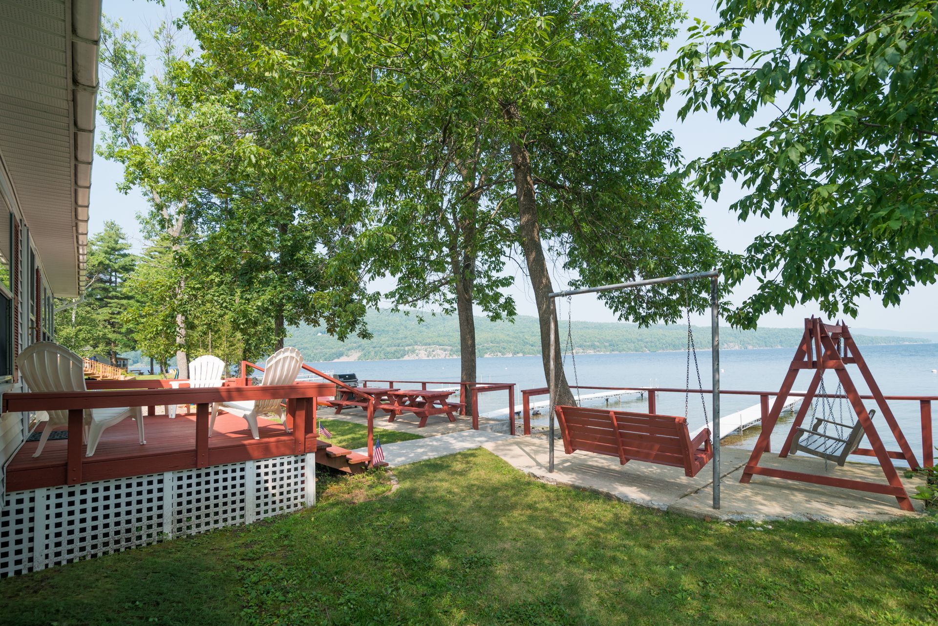 Lakeside deck with red chairs, picnic tables, and a swing under trees on a grassy lawn