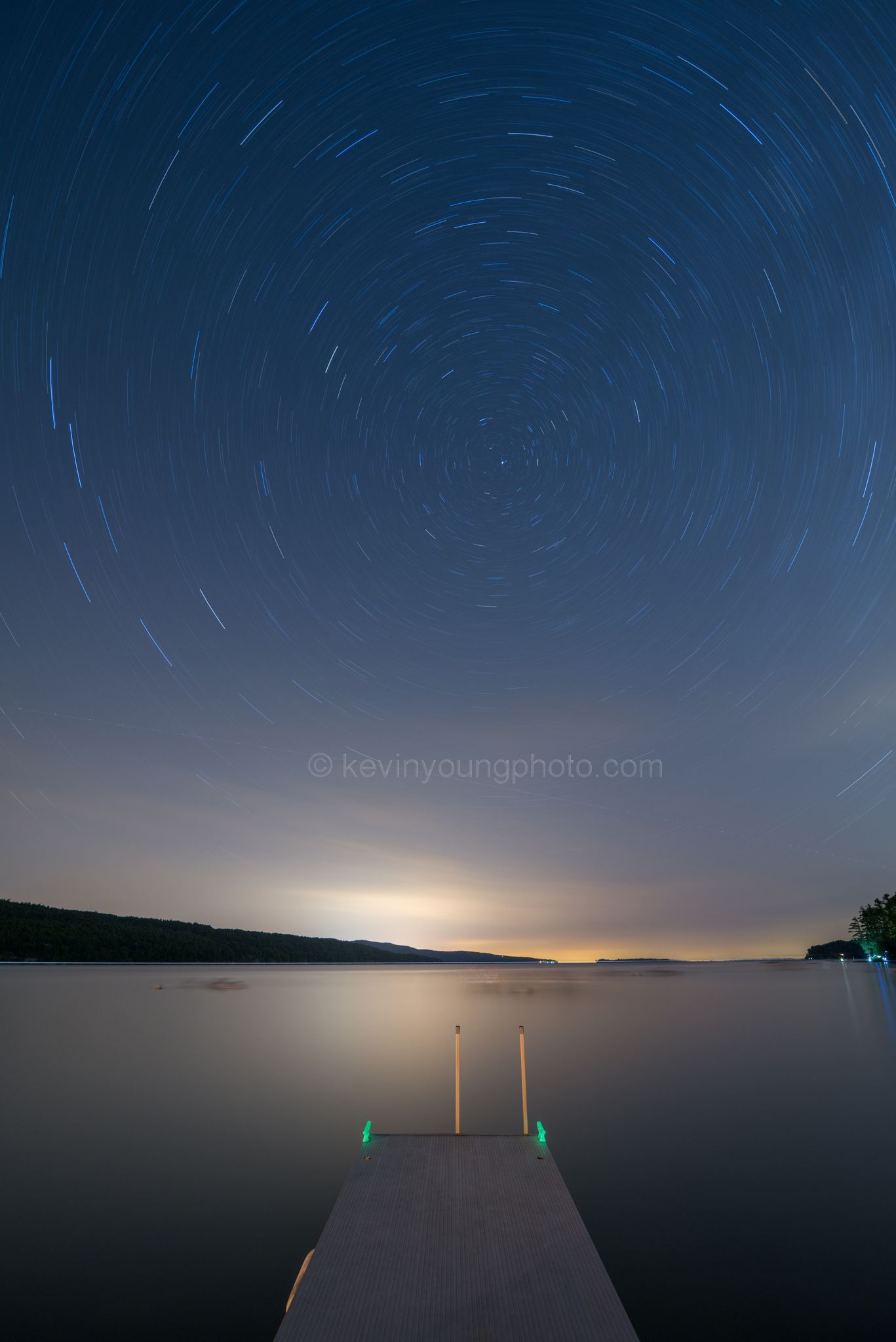 Night sky with star trails above a calm lake and a lit dock at dusk