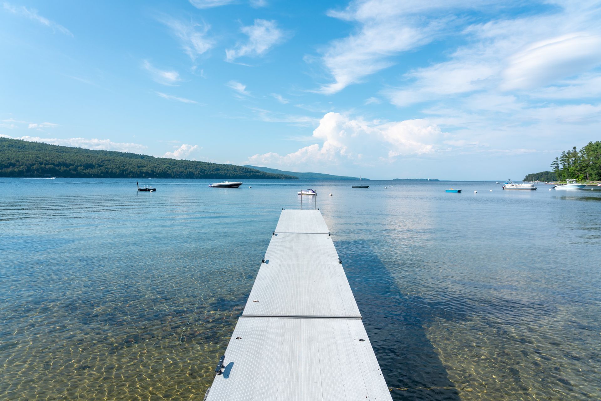 White dock stretching into a calm blue lake under a bright sky with distant hills and boats