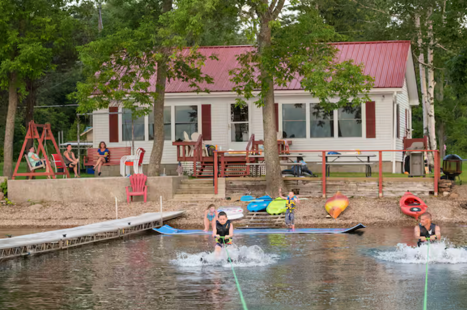 Children swimming and playing by a lakeside cabin with a red roof and dock.