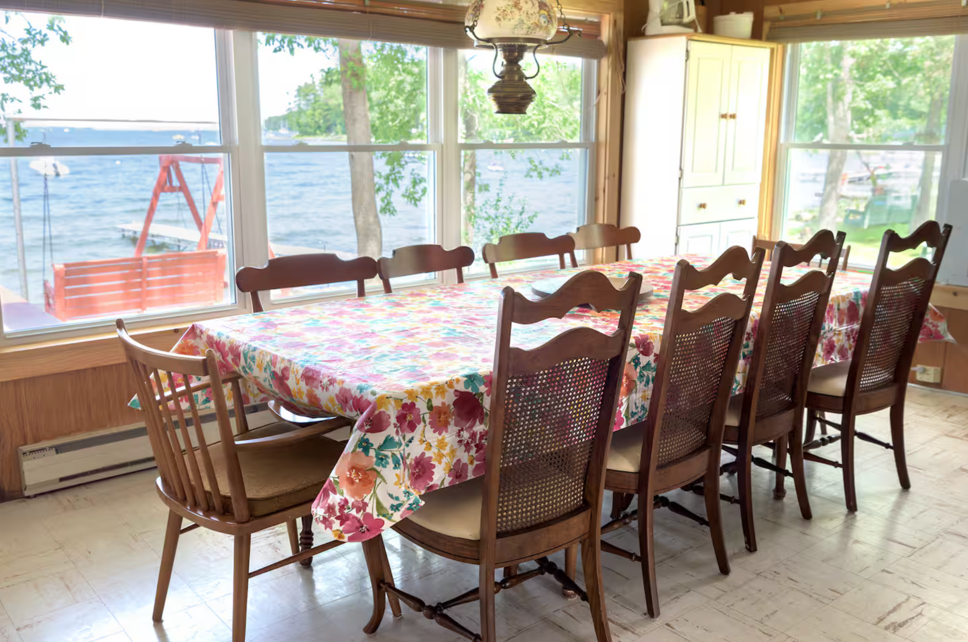 Dining room with long table, floral tablecloth, wooden chairs, and large windows overlooking the water