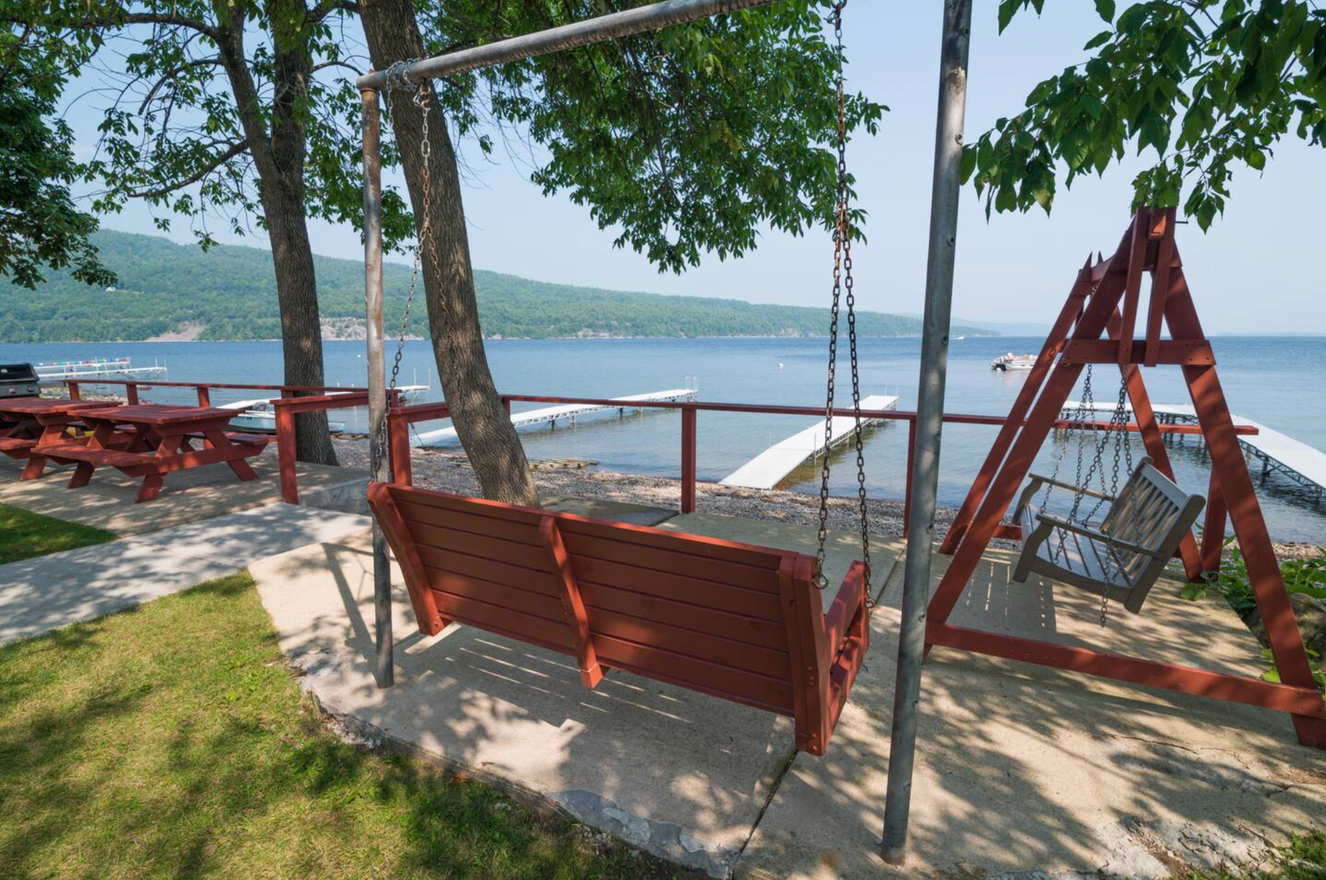 Red swings and picnic tables under trees on a lakeside patio with a view of the water