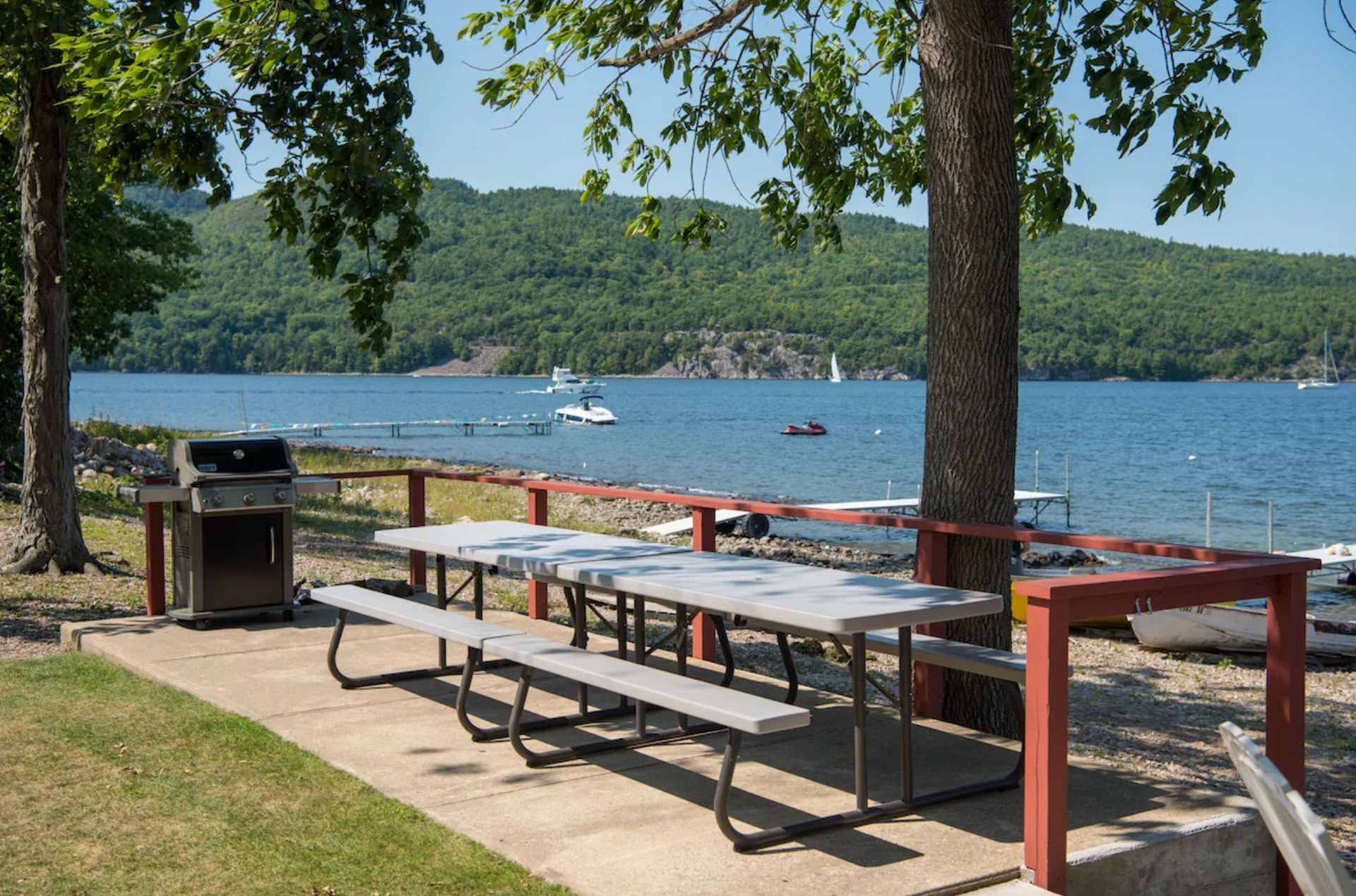 Picnic tables and a grill by a lakeside with trees and boats in the distance
