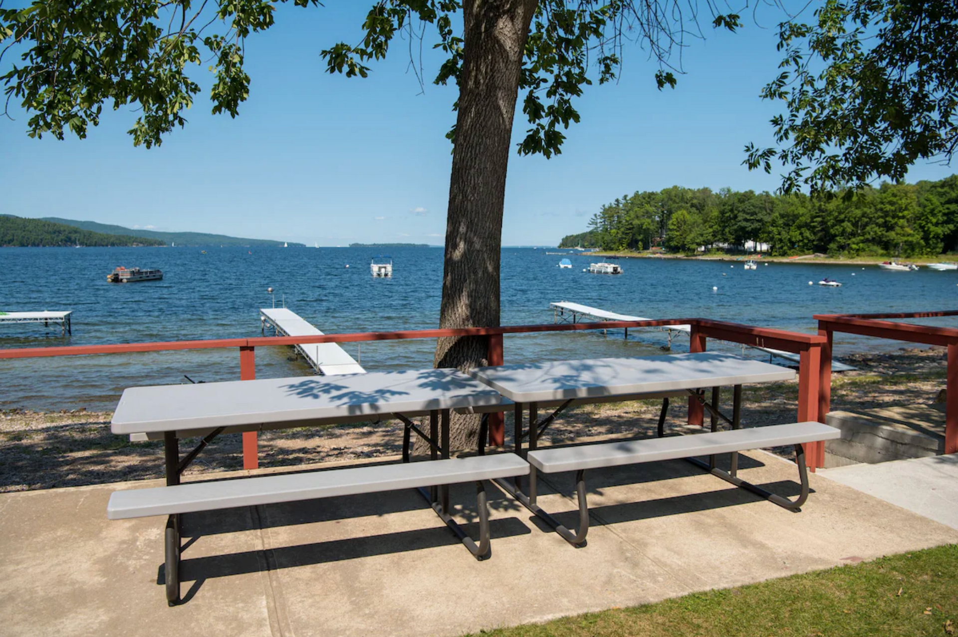 Picnic tables by a lakeside beach with a tree, dock railing, and boats on calm water