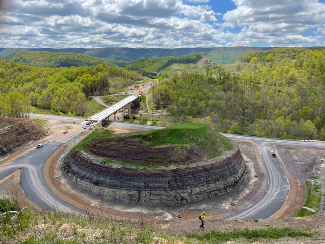 The image shows a highway construction project with forested hills in the background and a man in a safety vest in the foreground.