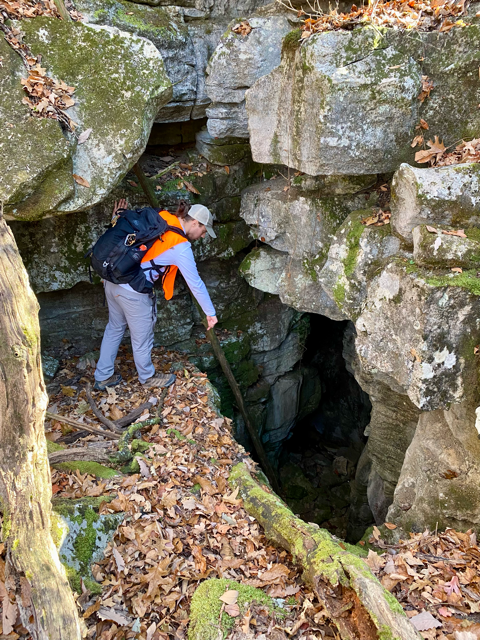 The image shows a man wearing a safety vest and backpack who is examining a cave entrance for bat habitat suitability.