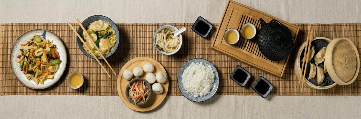 A top-down view of a traditional Chinese meal set on a bamboo mat, featuring various dishes, rice, dumplings, and tea.