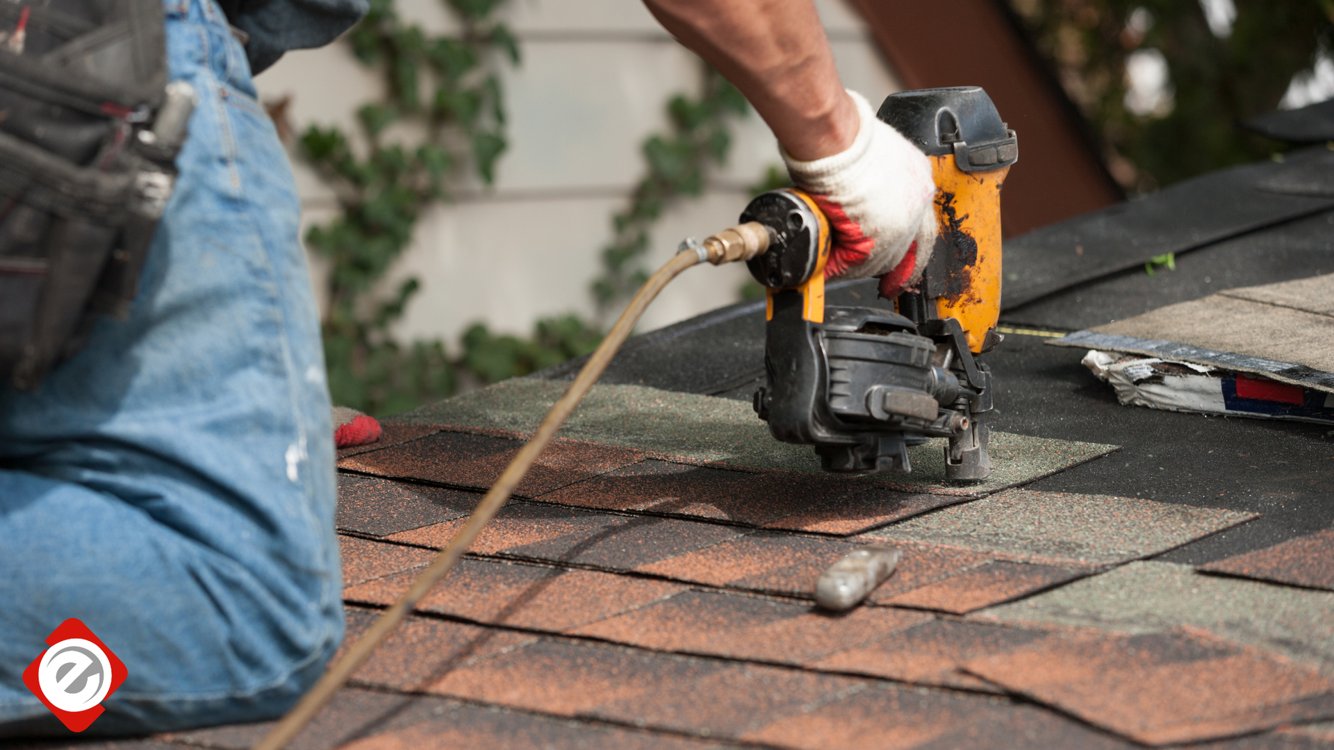 Roofer using a nail gun on a brown shingle roof, wearing gloves.