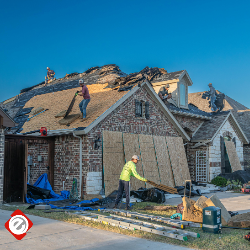Roofers replacing shingles on a residential home with brown brick and siding on a sunny day.