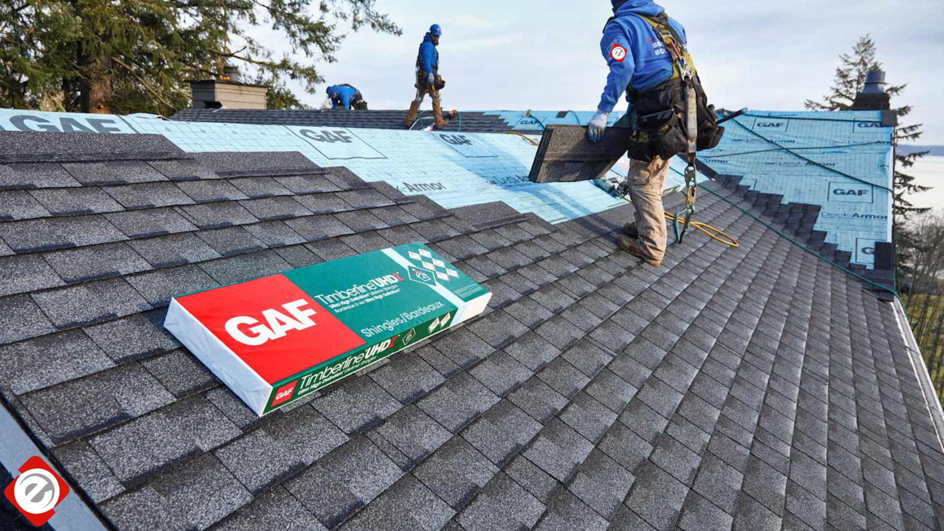 Roofers installing asphalt shingles on a residential roof, GAF shingle box in view.