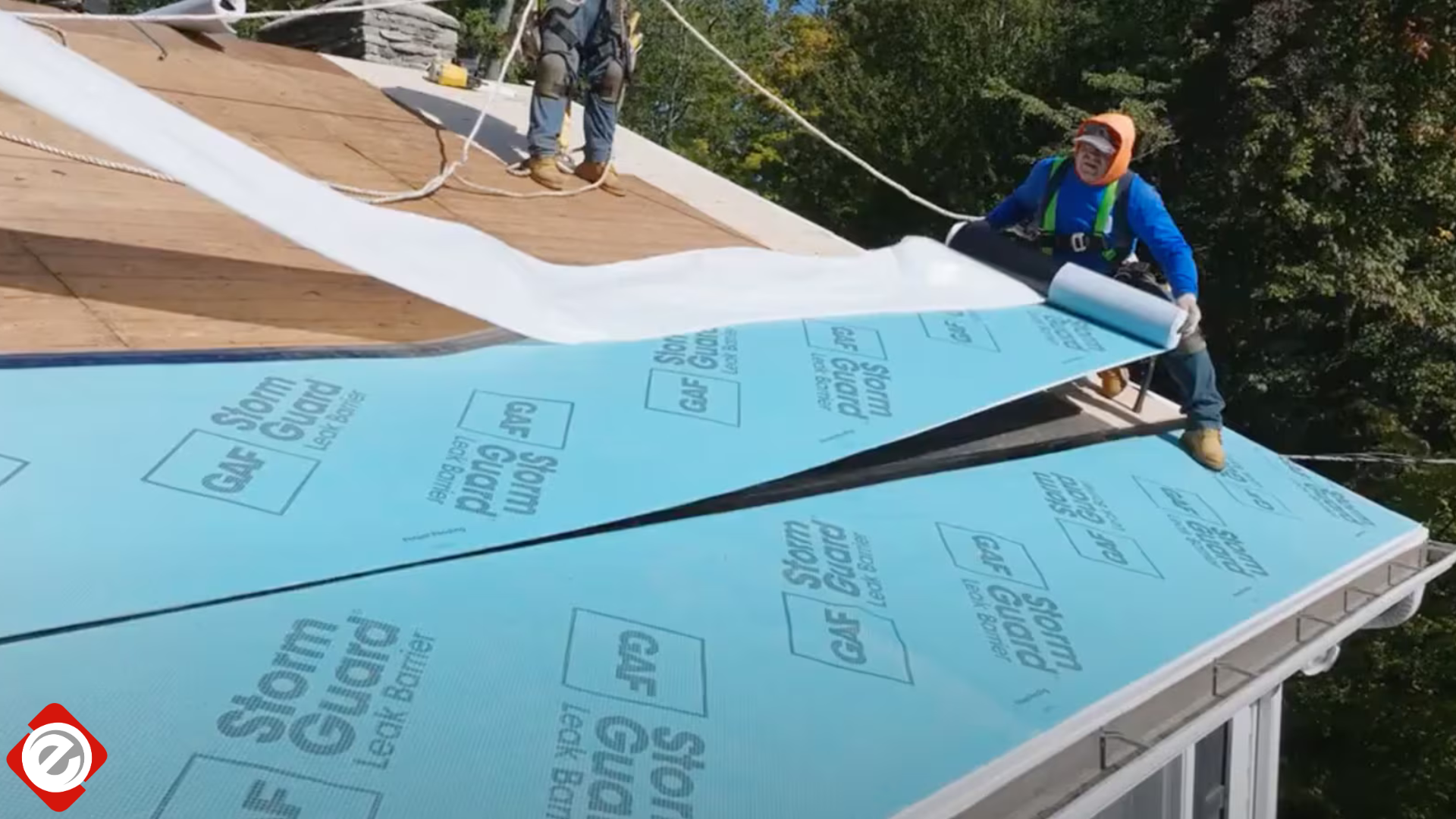 Workers installing roofing underlayment on a house roof. Blue material, safety harnesses.