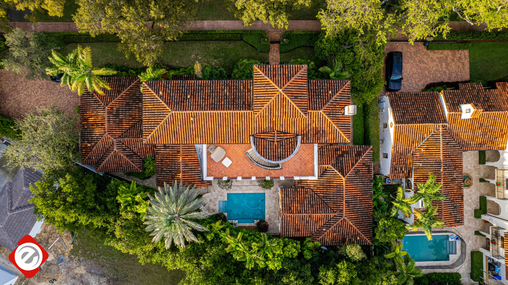 Overhead view of a large Spanish-style house with a red-tiled roof, a pool, and lush green landscaping.