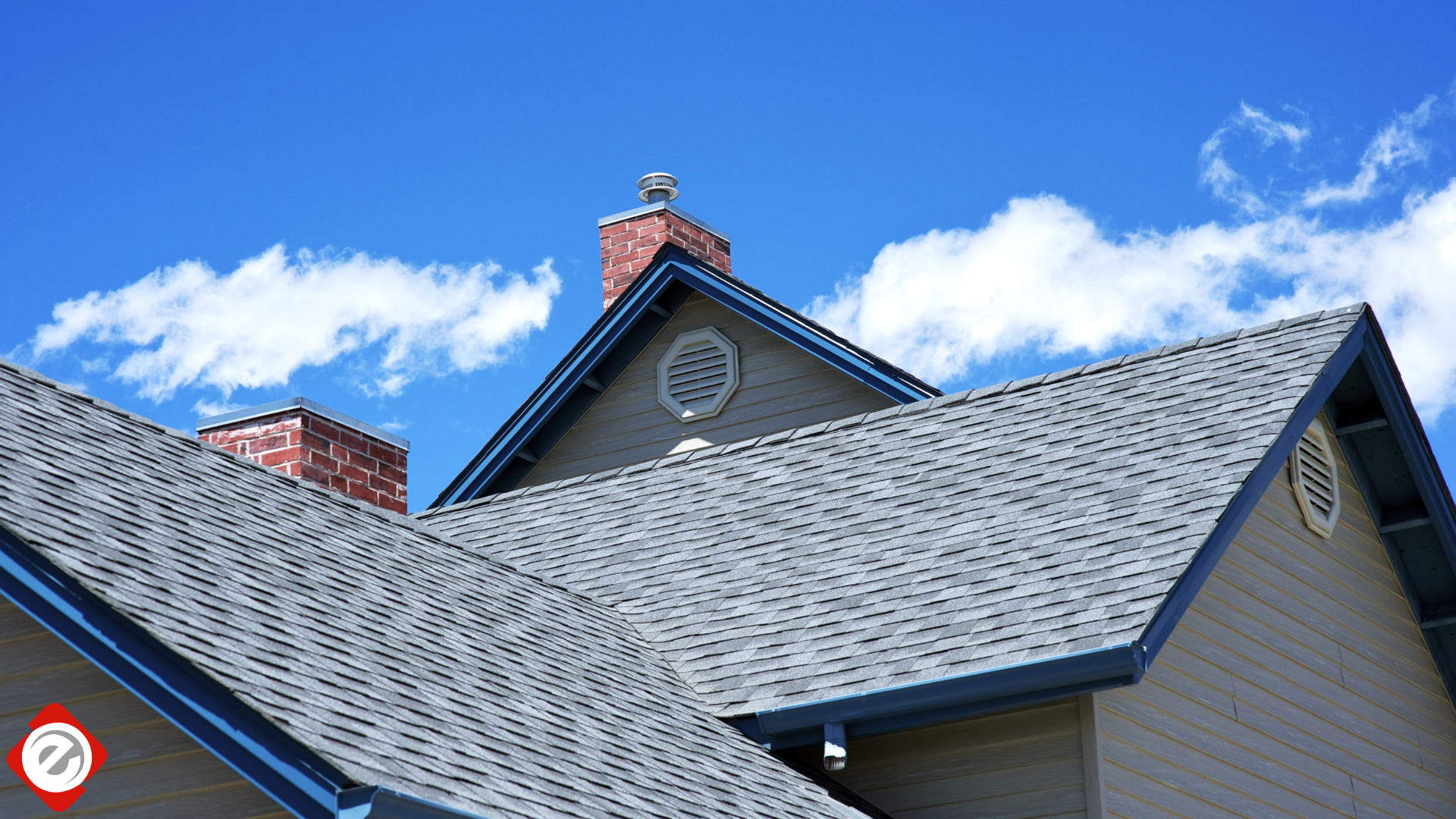 Gray shingled rooftops of a house against a blue sky with fluffy white clouds, with brick chimneys.