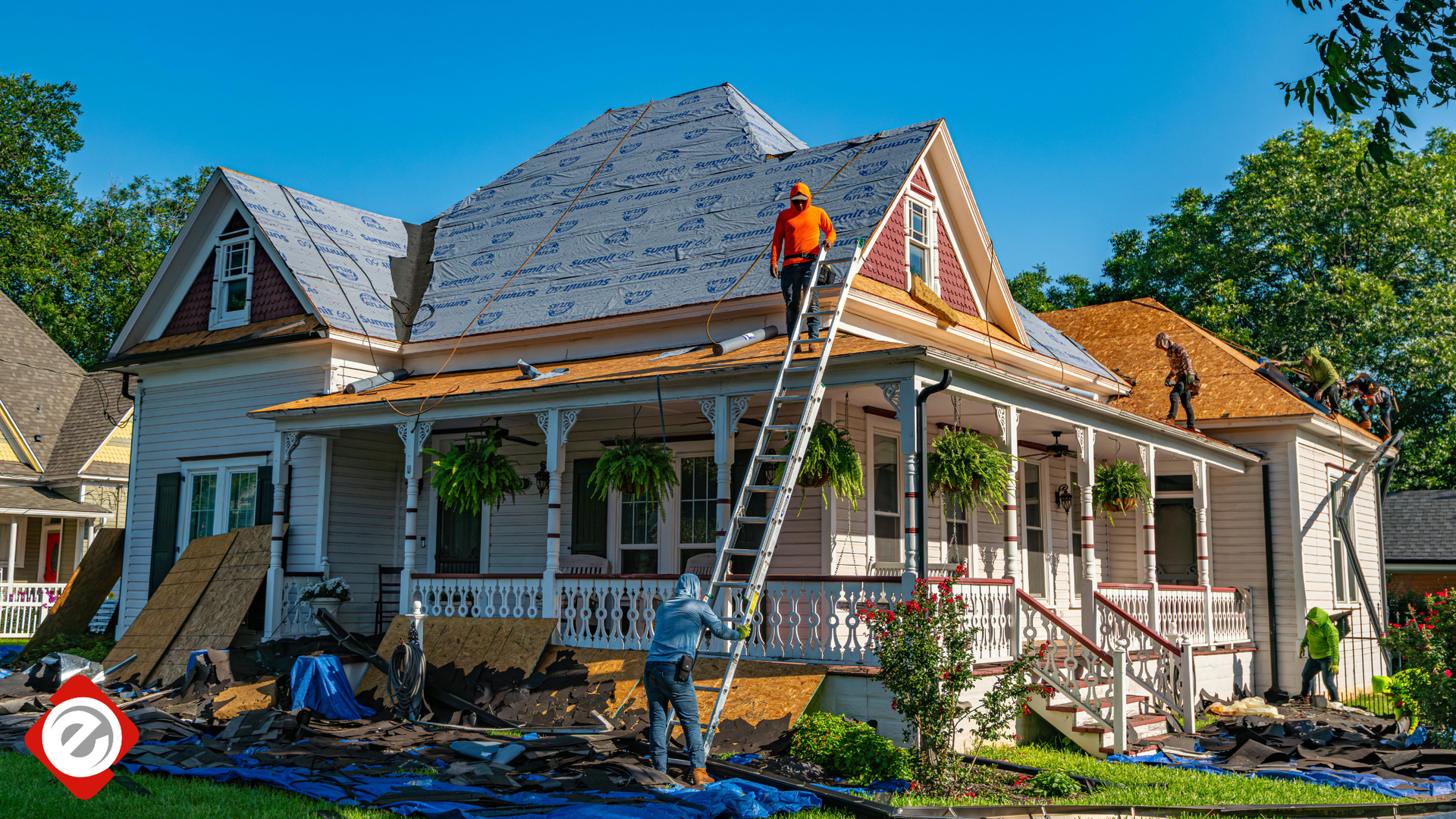 Workers replacing a roof on a two-story house. One on the roof, one climbing ladder, and another on the ground.
