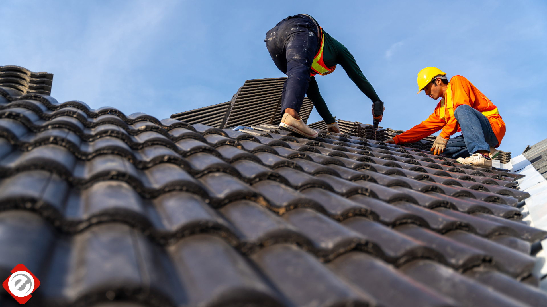 Two roofers installing dark ceramic roof tiles on a house under a blue sky.