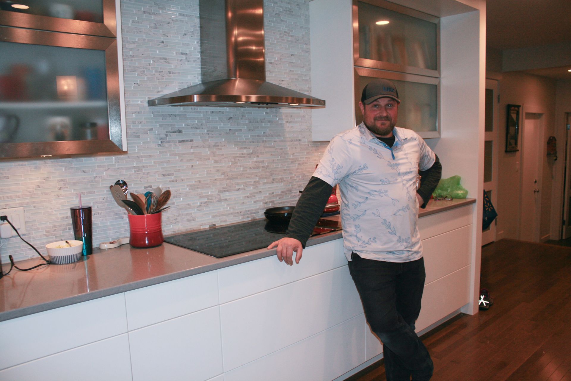 A person wearing a baseball cap stands in a modern kitchen, leaning against a white counter below a metal range hood.