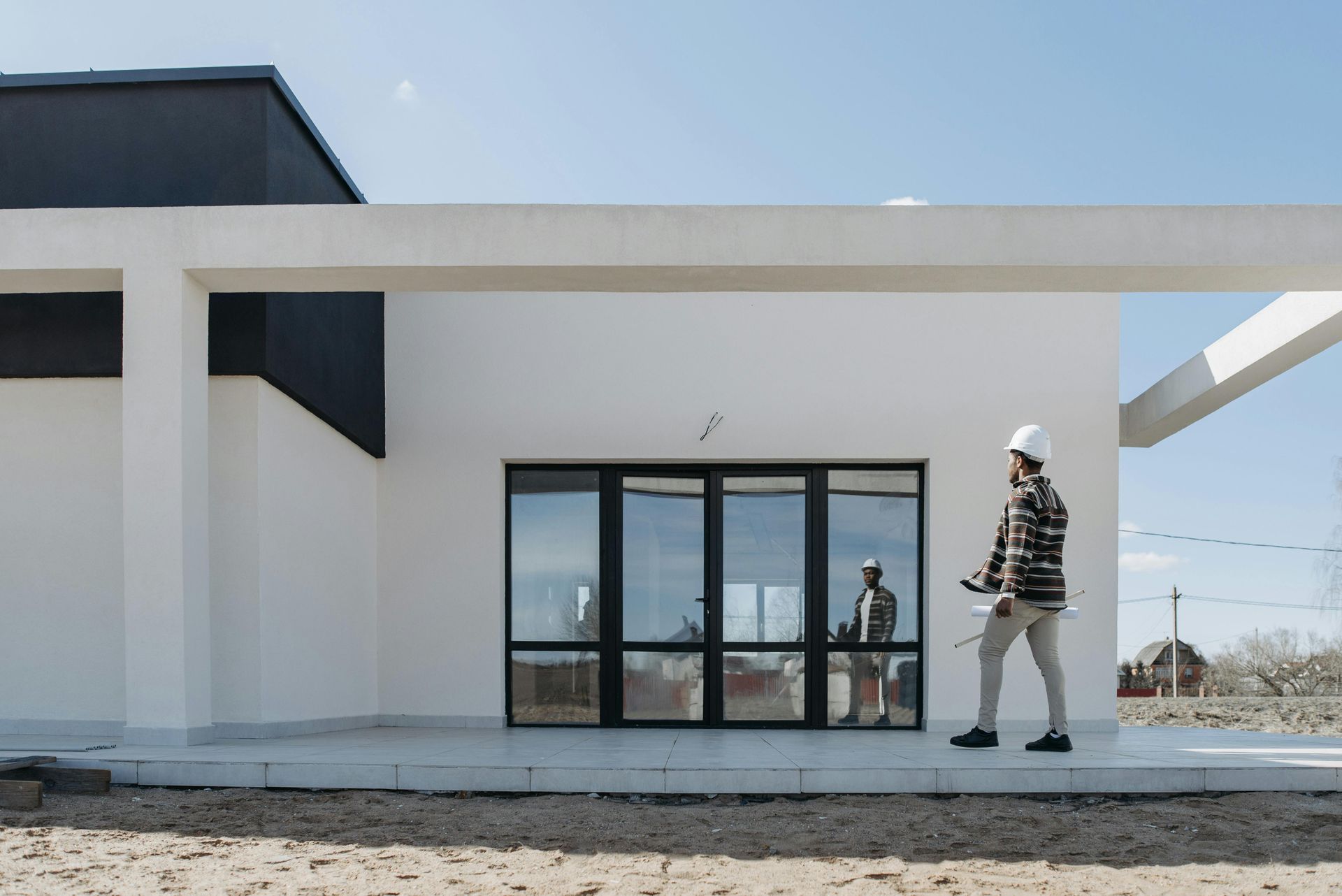 Architect in a hard hat inspecting a modern white building with large glass doors on a sunny day.