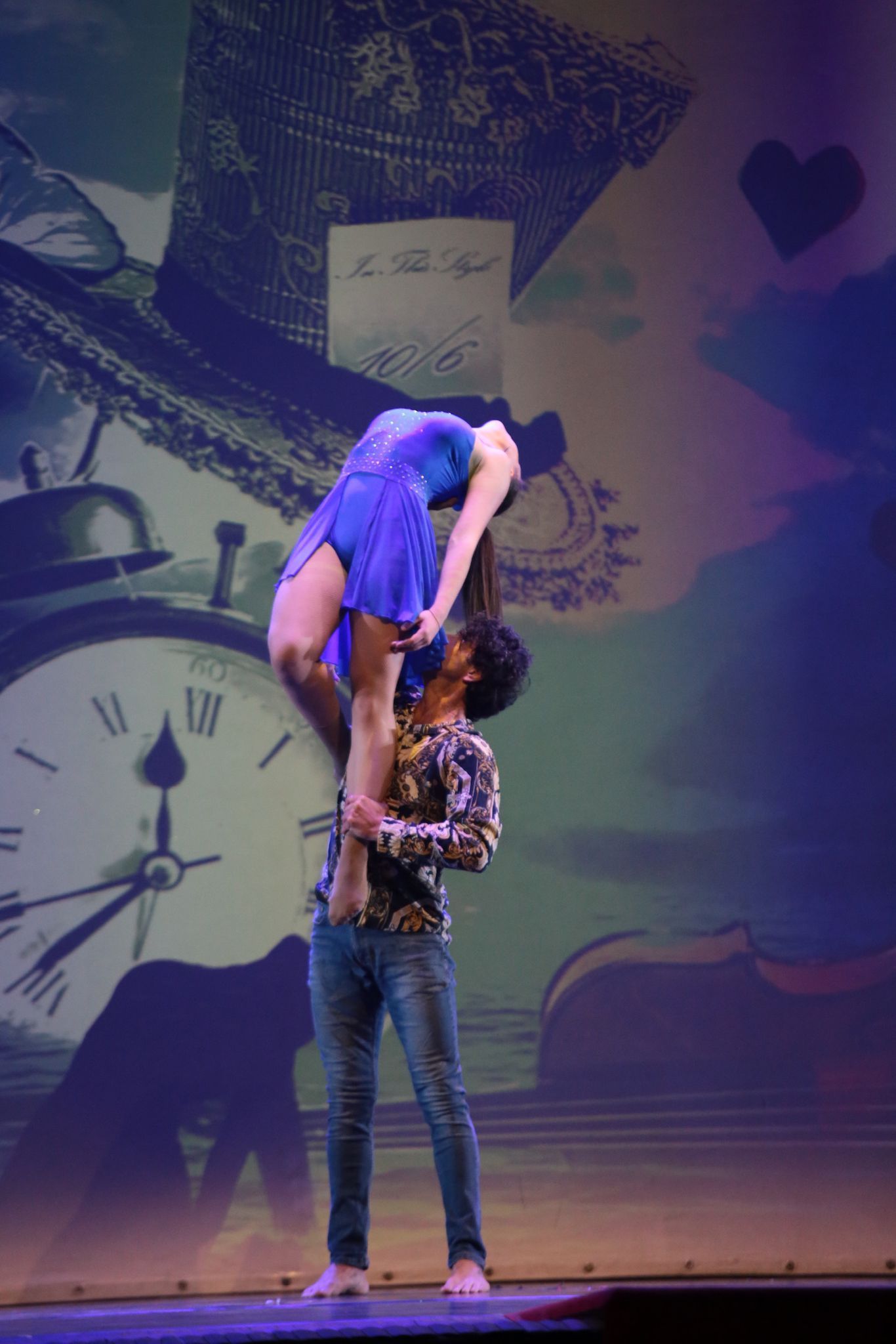 Un artista solleva un altro in una posa di danza davanti a uno sfondo che raffigura un orologio gigante e un cappello a cilindro di dimensioni spropositate.