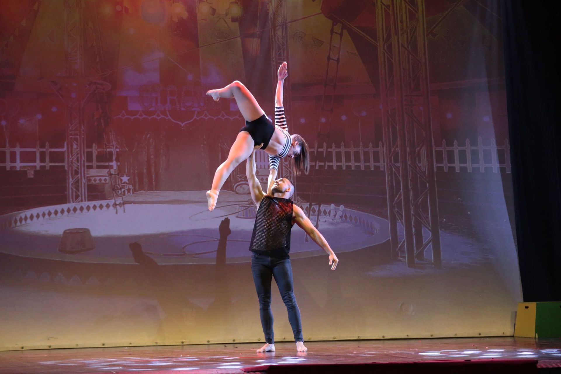 Durante uno spettacolo teatrale, un artista solleva un altro con una presa a una mano sopra la testa, sullo sfondo di una pista da circo.