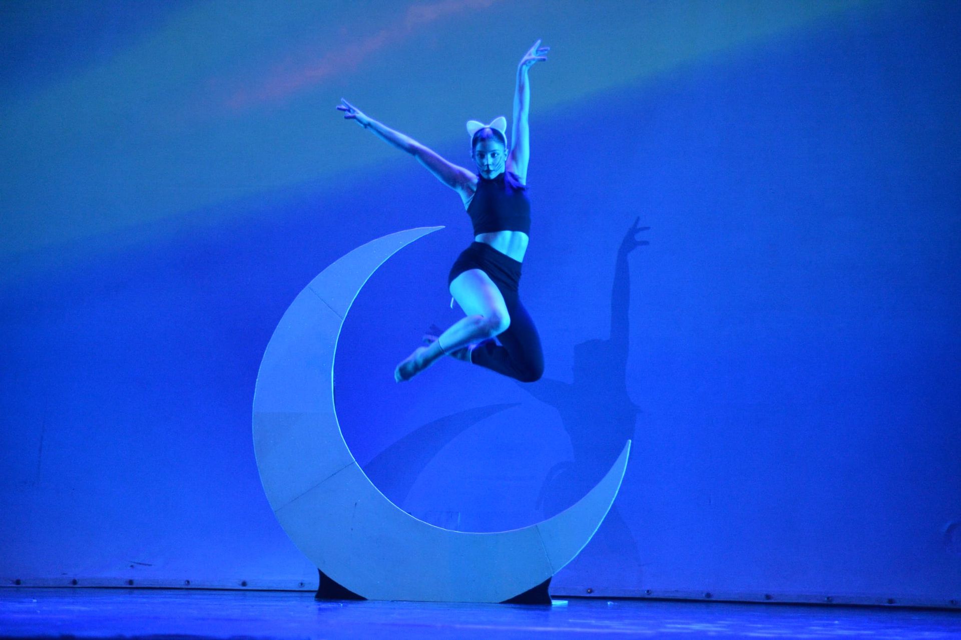 Una ballerina con un abito scuro salta davanti a una scenografia a forma di mezzaluna sullo sfondo di un palcoscenico illuminato di blu.